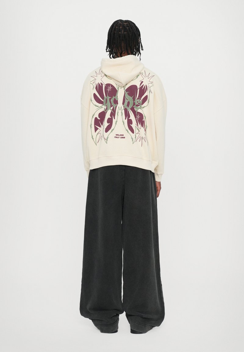 Cream hoodie with large burgundy graphic on back, featuring floral and thorn patterns. Paired with wide-leg black pants.