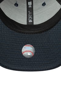 Tumši zilas tekstūras cepure ar rakstainu virsmu, centrā ar apļa MLB uzlīmi, uz kuras attēlots beisbola grafiks un teksts.