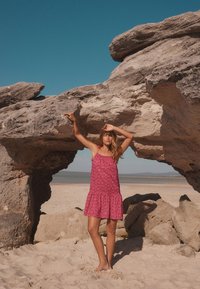 Vestito rosa con fantasia, orlo svasato e spalline, indossato da una persona in piedi sotto una grande formazione rocciosa su una spiaggia sabbiosa.