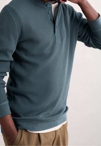 Top a maniche lunghe in stile Henley in tessuto a coste color teal, con un'apertura a tre bottoni e polsini a contrasto. Indossato sopra una camicia bianca.