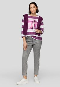 Lässiges, langärmeliges Oberteil mit lila und weißen Streifen, das ein grafisches Design aufweist. Kombiniert mit grauen Jeans und weißen Turnschuhen.