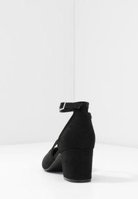 Talón de ante negro con una correa, mostrando un diseño afilado. Posicionado sobre un fondo blanco liso, enfatizando el elegante perfil del zapato.