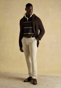 Barna bordázott kardigán egy sötétkék csíkos póló felett, amely zöld és sárga csíkokkal díszített, krémszínű nadrág, és barna loaferek kontrasztos részletekkel.