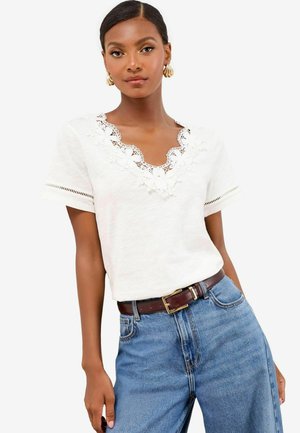 Woman wearing white short-sleeve top with floral lace V-neck detail, blue jeans, brown belt, and gold shell-shaped earrings on white background.