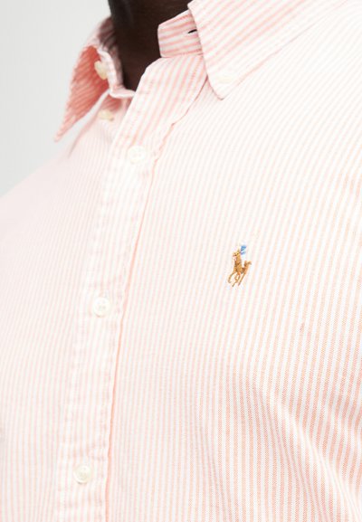Zbliżenie na osobę w jasnoróżowej koszuli w białe paski z guzikami, z małym haftowanym logiem gracza polo.