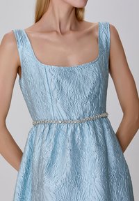 Robe bleu clair avec un tissu texturé, décolleté carré et taille froncée. Présente une ceinture en argent perlée pour l'accentuation et la structure.