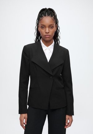 Femme aux cheveux tressés portant un blazer noir cintré et une chemise blanche, debout devant un fond clair uni, faisant face à l’avant.