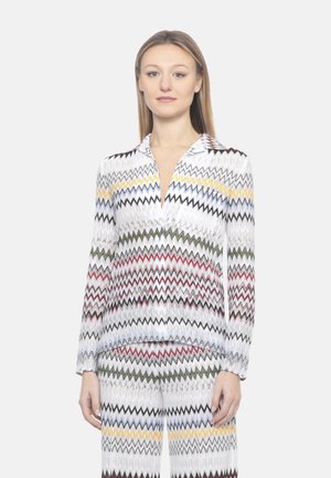 Donna in piedi che indossa un top bianco a maniche lunghe e pantaloni con motivo a zigzag multicolore su uno sfondo chiaro e uniforme.