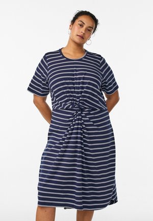 Woman with dark hair in a navy blue dress with white horizontal stripes and a front twist detail, standing against a plain light background.