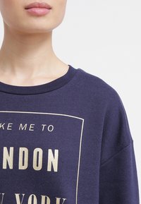 Felpa navy con scollatura rotonda e maniche corte, caratterizzata da una stampa grafica dorata che include il testo "LONDON" e "NEW YORK."