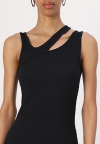 Svart ribbet tanktopp med en enkelt skulderstropp og et utskjæringselement. Stoffet er glatt med en tettsittende design og teksturert overflate.