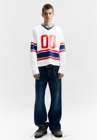 Fehér hosszú ujjú ing piros és kék csíkokkal, nagy piros "00" grafikával. Laza kék farmernadrággal és szürke lábbelivel viselve.