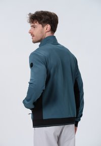 Giacca sportiva da uomo con parte superiore testurizzata turchese, sezione inferiore nera, colletto alto e dettagli con zip.