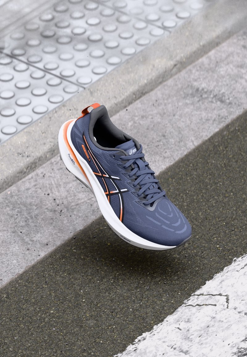 Chaussure de course bleue et orange en l'air au-dessus d'un trottoir texturé et d'une rue asphaltée avec marquage blanc.
