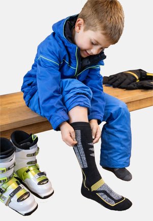 Black ski socks with gray and yellow stripes, featuring a cushioned design. A child wearing a blue jacket sits on a wooden bench with ski boots.
