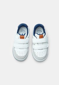 Baskets blanches avec deux bandes Velcro, accents bleus et un détail beige. Le matériau est synthétique, avec une pointe perforée et des bordures grises.