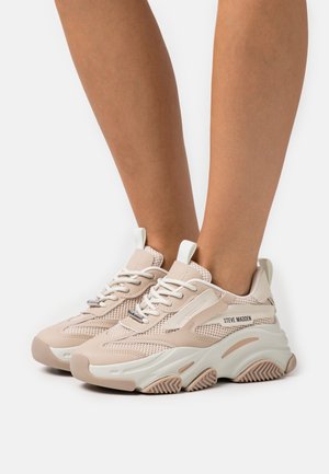 Beżowe siateczkowe i skórzane sneakersy z białymi akcentami, okrągłym noskiem, grubą podeszwą i wyraźnym wzorem bieżnika. Nazwa marki "Steve Madden" widoczna z boku.