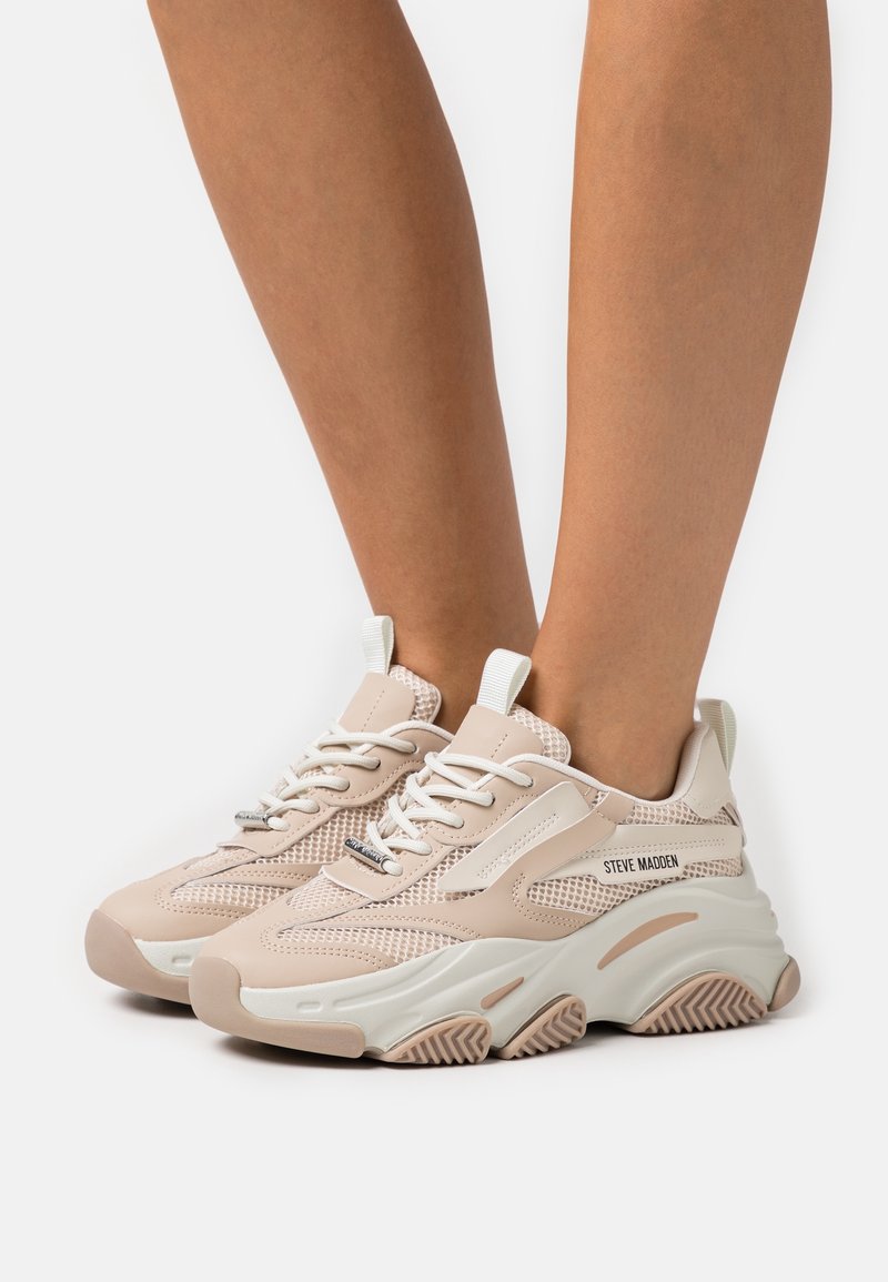 Zapatillas de malla y cuero beige con acentos blancos, punta redondeada, suela gruesa y un patrón de banda de rodadura prominente. Nombre de la marca "Steve Madden" visible en el costado.