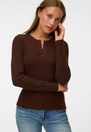 Femme à la peau claire et aux cheveux châtain clair portant un haut marron côtelé à manches longues et un jean bleu, posant avec la main près du visage sur fond blanc.