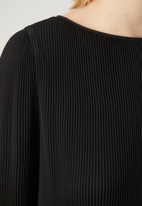 Haut plissé noir avec un col rond et un tissu texturé à côtes verticales, mettant en valeur le contraste entre la lumière et l'ombre.