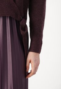 Mörkröd stickad tröja med ribbad textur, med en sidoförslutning. Kombinerad med en plisserad, ljuslila kjol med vertikala ränder.