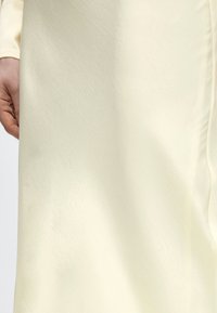 Χέρι που ακουμπά δίπλα σε υφή υφάσματος ανοιχτού κρεμ χρώματος με λεπτό διαγώνιο μοτίβο και απαλά διπλώματα ορατά στη δεξιά πλευρά.