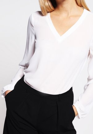 Blusa bianca a maniche lunghe con scollo a V, realizzata in tessuto morbido e leggero, con maniche leggermente affusolate; abbinata a pantaloni neri su misura.