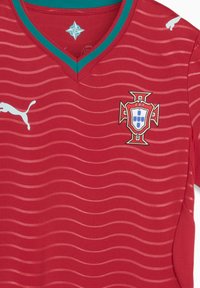 Κόκκινη φανέλα ποδοσφαίρου με κυματοειδές μοτίβο, V-λαιμόκοψη, στοιχείο σε τιρκουάζ και λογότυπο Puma στο αριστερό στήθος. Εμφανίζεται το έμβλημα της Ομοσπονδίας Ποδοσφαίρου Πορτογαλίας.