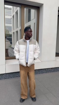 Puffy white and beige jacket with a high collar, worn over a light t-shirt. Tan wide-leg pants and black boots. Standing against a modern building.