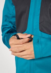 Miętowa kurtka z czarną górną częścią, z zamkiem błyskawicznym z przodu i otworami na kciuki w rękawach. Gładka faktura i sportowy krój.