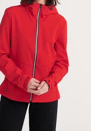 Person trägt eine leuchtend rote Jacke mit Reißverschluss und Kapuze, steht mit verschränkten Händen, bekleidet mit schwarzen Hosen vor einfarbigem Hintergrund.
