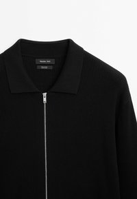 Schwarzes Baumwollshirt mit Reißverschluss und geripptem Muster, das einen umgefalteten Kragen und silberne Reißverschlussdetails an der Vorderseite aufweist.