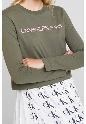 Nainen, joka on ristissä käsivarret päällään, pukeutuneena oliivinvihreään pitkähihnaiseen yläosaan, jossa lukee "Calvin Klein Jeans" ja valkoiseen hameeseen, jossa on toistuva CK-logo.