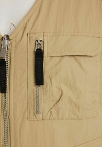 Beež kerge vest, millel on ees hõbedane tõmblukk, must tõmbluku tasku ja lapp-tasks. Materjalil on kergelt kortsus tekstuur.