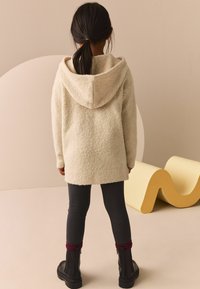 Un pull à capuche texturé de couleur crème, présenté de dos, associé à un legging gris foncé et des bottes noires, sur un fond minimal.
