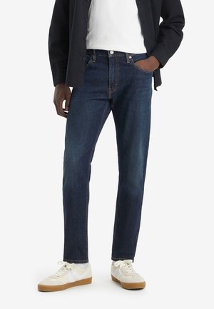 Person trägt dunkelblaue Jeans, weiße Sneaker, weißes T-Shirt und schwarze Jacke, eine Hand in der Tasche, steht vor weißem Hintergrund.