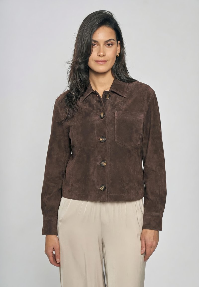 Donna con capelli lunghi e scuri che indossa una giacca di pelle scamosciata marrone con bottoni e pantaloni beige chiaro su uno sfondo grigio uniforme.