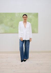 Femme portant un blazer blanc texturé, un jean bleu à jambes larges et des talons noirs pointus, debout à l'intérieur devant une œuvre d'art murale abstraite verte.