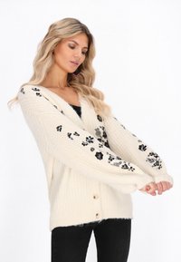 Crèmekleurige gebreide cardigan met lange mouwen, V-hals en zwart florale versieringen. Kenmerken zijn houten knopen en een zachte, gestructureerde stof.
