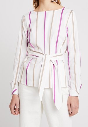 Valkoinen pitkähihainen pusero, jossa pystysuorat beige ja vaaleanpunaiset raidat, vyötäröllä solmittu vyö, yhdistetty valkoisiin housuihin.