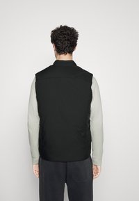 Svart ermeløs vest med en jevn tekstur, båret over en lys grå langermet skjorte. Minimalistisk design med høy krage og ingen synlig maskinvare.