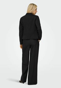 Femme aux longs cheveux blonds portant un blazer noir, un pantalon noir à jambes larges et des talons noirs, debout de dos avec une main dans la poche.