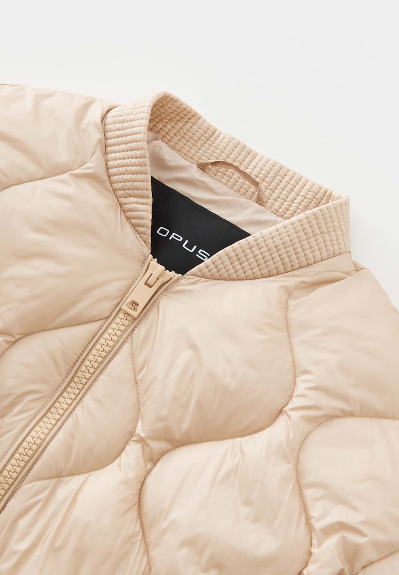 Helles beiges gesteppter Jacke mit einem strukturierten Rippkragen und einem Frontreißverschluss. Das Innenetikett trägt die Aufschrift "OPUS." Glatte, glänzende Stoffoberfläche.
