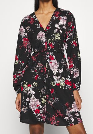 Musta kukkakuosinen mekko, jossa on pitkät hihat, V-aukko ja vyötärösolki. Mekossa on värikkäitä kukkakuvioita punaisena, vaaleanpunaisena ja vihreänä.