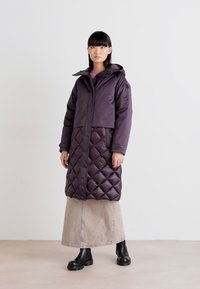 Palton izolat de culoare purpurie, cu o secțiune inferioară matlasată, glugă și fermoar. Modelul poartă o fustă gri din denim și cizme negre. Textură netedă și lucioasă.