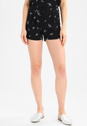 Kvinde iført sorte shorts med hvide fuglemønstre og hvide slip-on sko, stående mod en ensfarvet lys baggrund.