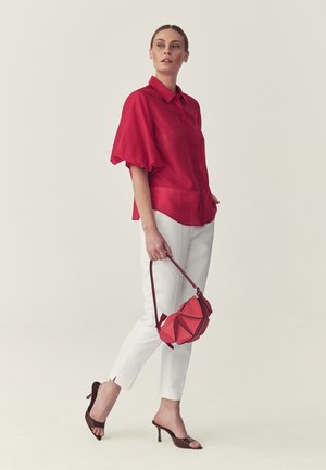 Mujer de pie con una blusa roja, pantalones blancos, sandalias marrones con tacón y sosteniendo un bolso rosa contra un fondo liso y claro.