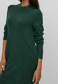 Abito verde lavorato a maglia con scollatura alta, maniche lunghe e polsini a costine. Tessuto morbido e silhouette aderente, che arriva a metà polpaccio.