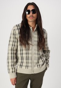 Beige strikket cardigan med svart og oliven grønn rutemønster, frontknapper, ribbede mansjetter, båret over en lys skjorte og brune bukser.