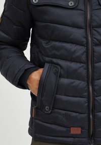 Marineblå pufferjakke med quiltet design, glidelås, trykknapper og lærlogo. Har ribbestrikkede mansjetter og sidelommer.